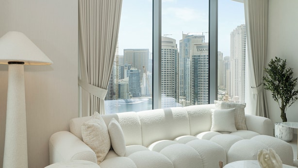 A modern living room with a large window, a plush white sofa, a round marble coffee table, and a floor lamp. - Experience 2BR Luxury Living  LIV Marina (Dubai)