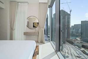 A modern hotel room with a large window, a bed, a round mirror, and a view of a cityscape with construction cranes. - Experience 2BR Luxury Living  LIV Marina (Dubai)
