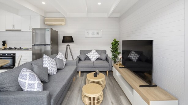 Ein modernes Wohnzimmer mit einem grauen Sofa, zwei runden Holztischen, einem Fernseher und einer Küchenzeile mit weißen Schränken und einem Kühlschrank.