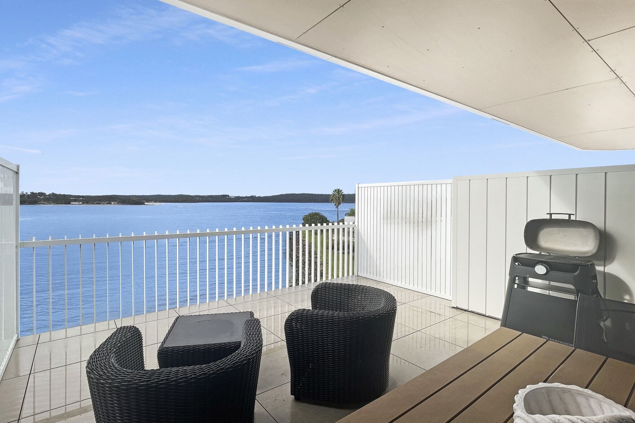 A balcony with wicker furniture, a grill, and a view of a calm body of water.