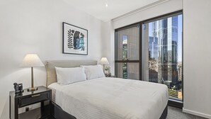 A modern hotel room with a large bed, two bedside lamps, a framed artwork, and a view of a cityscape through floor-to-ceiling windows. - Elegant Southbank Escape Apartment with Balcony (Southbank)