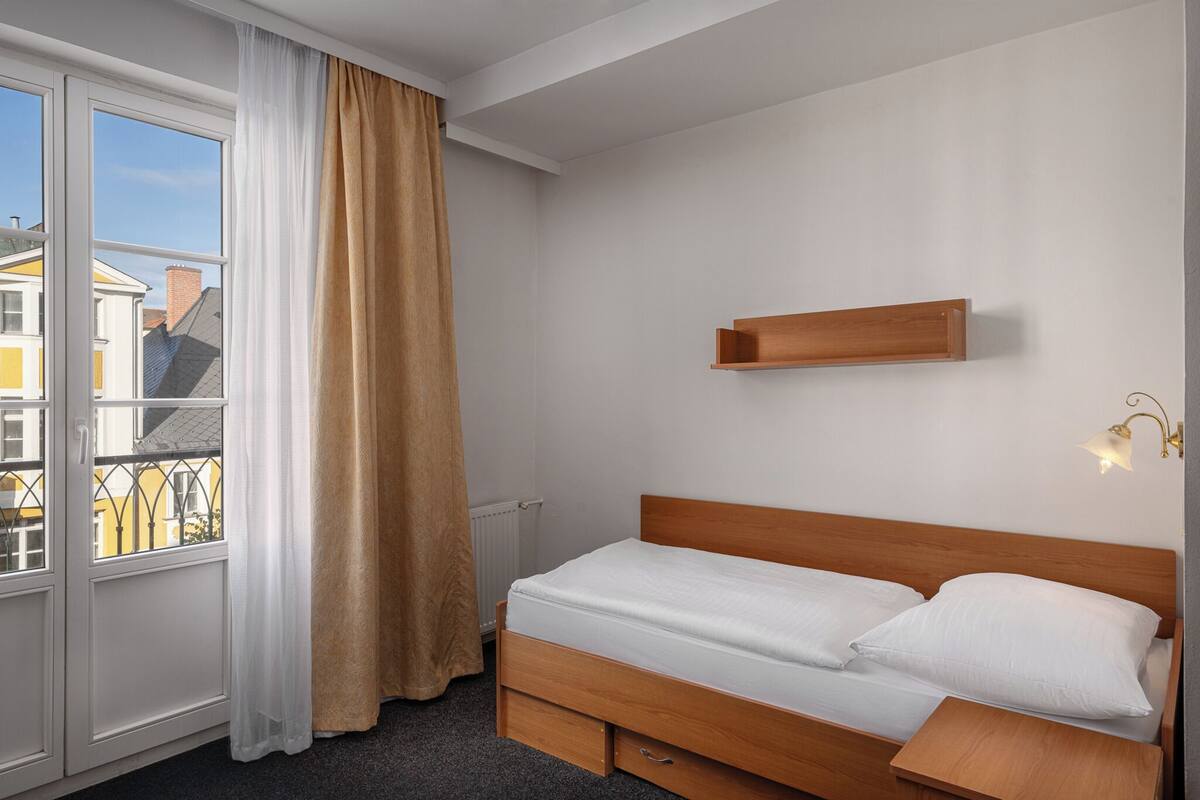 Una cama individual con cabecera de madera, una mesita de noche, un pequeño estante sobre la cama y una ventana con cortina.