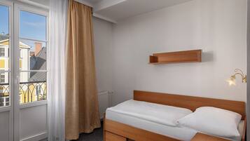 A single bed with a wooden headboard, a bedside table, a small shelf above the bed, and a window with a curtain.