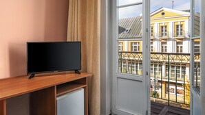 A room with a television on a wooden stand, a window with a view of a yellow building, and a door with a balcony. - Hotel Goethe Spa & Medical (Františkovy Lázně)