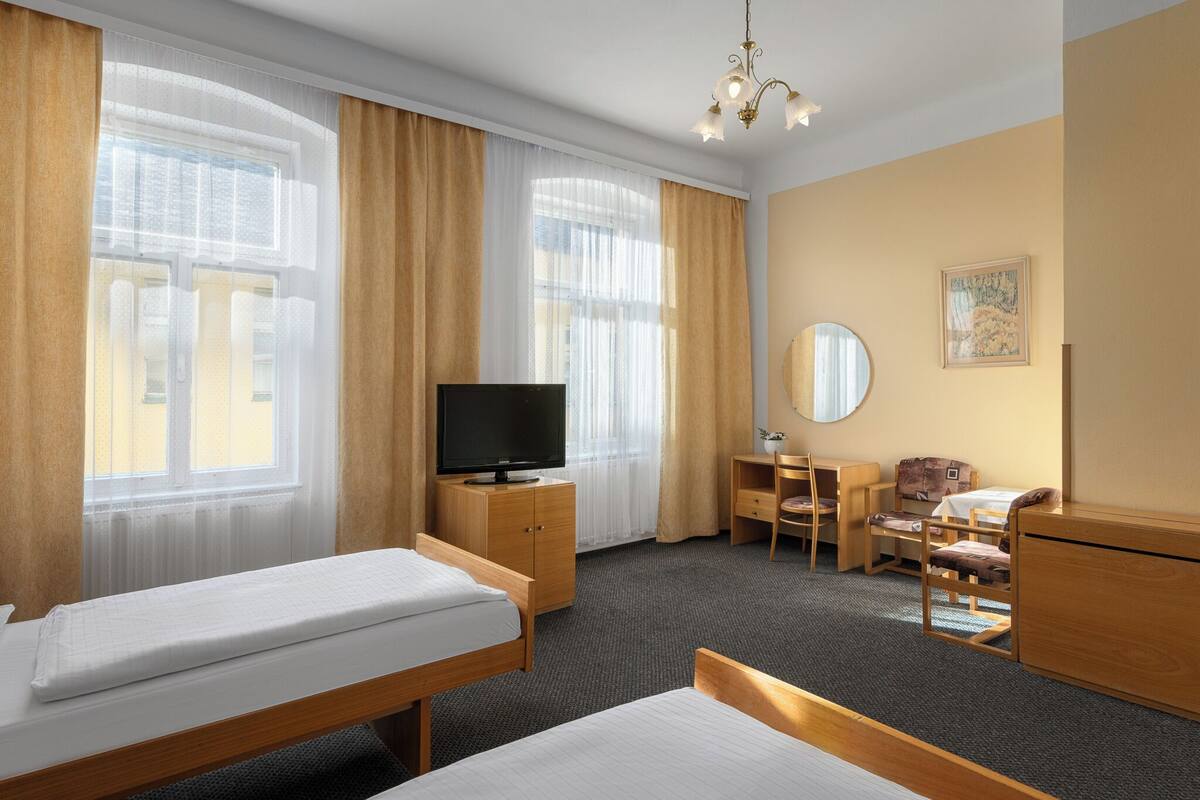 Habitación de hotel con dos camas, un escritorio, una silla, televisión y ventanas amplias con cortinas.