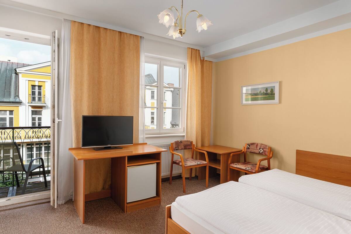 Habitación de hotel con cama, escritorio con televisor, silla, mueble bajo y ventana con cortinas.