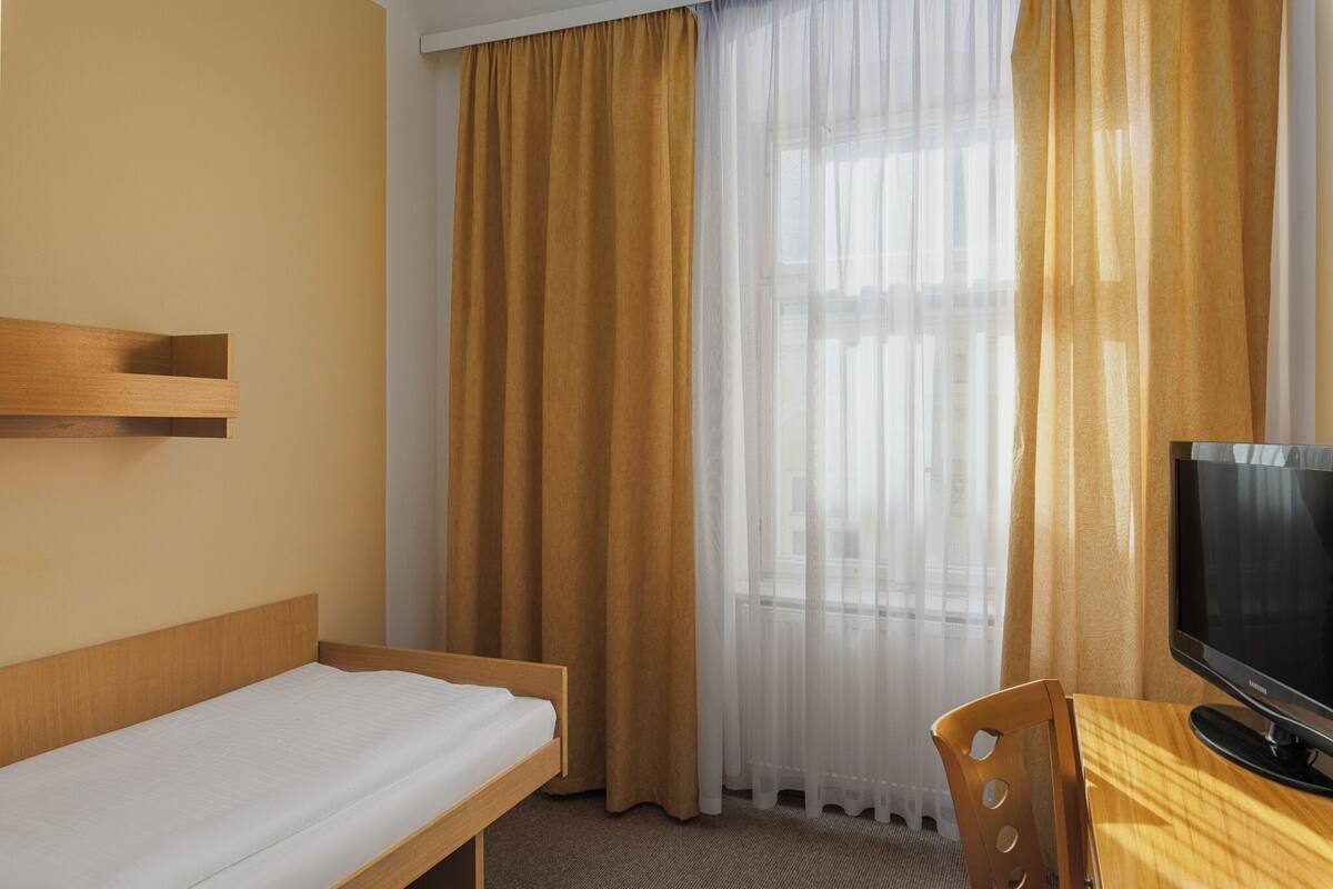 Una habitación de hotel con una cama individual, un escritorio de madera y cortinas translúcidas.