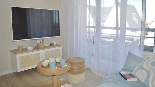 A modern living room with a flat-screen TV, a wooden console table with cups and a basket, a round wooden stool, and a sofa with a book. - La parenthèse bleue, loggia and parking lot (Courseulles-sur-Mer)