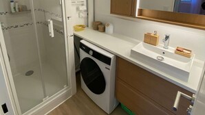 A compact bathroom with a glass shower enclosure, a washing machine, a sink with a mirror, and wooden cabinets. - La parenthèse bleue, loggia and parking lot (Courseulles-sur-Mer)