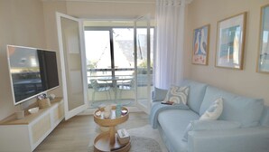 A modern living room with a flat-screen TV, a sofa, a coffee table, and a balcony with a table and chairs. - La parenthèse bleue, loggia and parking lot (Courseulles-sur-Mer)