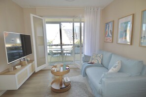 A modern living room with a flat-screen TV, a sofa, a coffee table, and a balcony with a table and chairs. - La parenthèse bleue, loggia and parking lot (Courseulles-sur-Mer)
