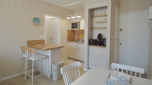 A compact kitchen with a microwave, coffee maker, and shelves with glasses. There is a dining area with a table and chairs. - La parenthèse bleue, loggia and parking lot (Courseulles-sur-Mer)