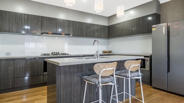 A modern kitchen with dark wood cabinetry, a central island with bar stools, stainless steel appliances, and pendant lighting. - Spacious 4-Bed House with Outdoor Area (Byford)