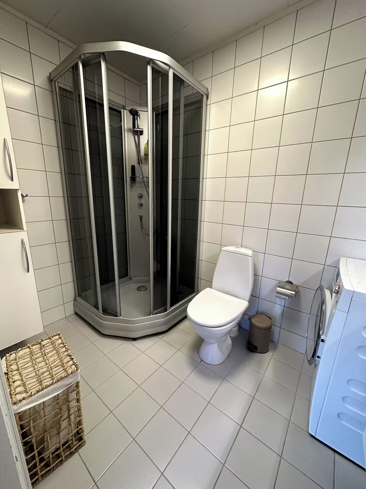 A bathroom with a glass-enclosed shower, a toilet, a trash bin, and a washing machine.