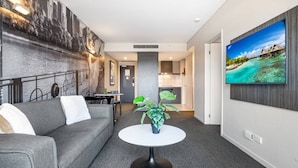 Ein modernes Wohnzimmer mit einem grauen Sofa, einem runden weißen Tisch mit einer Pflanze und einem großen Fernseher an der Wand, der eine tropische Strandlandschaft zeigt.