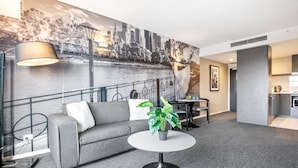 Ein modernes Hotelzimmer mit grauem Sofa, einem runden Tisch mit Pflanze, einer Stadtlandschaftsmalerei, einem Essbereich und einer Küchenzeile.