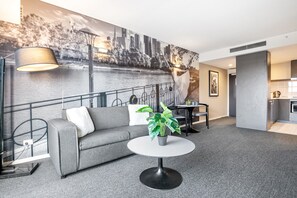 A modern hotel room with a grey sofa, a round table with a plant, a cityscape mural, a dining area, and a kitchenette. - Spacious Apartment in Heart of Southbank (Southbank)