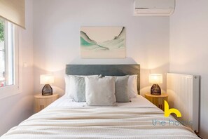 Une chambre à coucher bien rangée, avec un lit, deux tables de chevet, un climatiseur fixé au mur et un tableau au-dessus du lit.