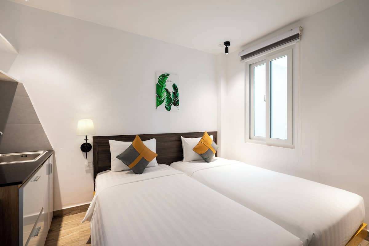 Habitación de hotel con dos camas, una kitchenette pequeña, una obra de arte colgada en la pared y una ventana con persianas.