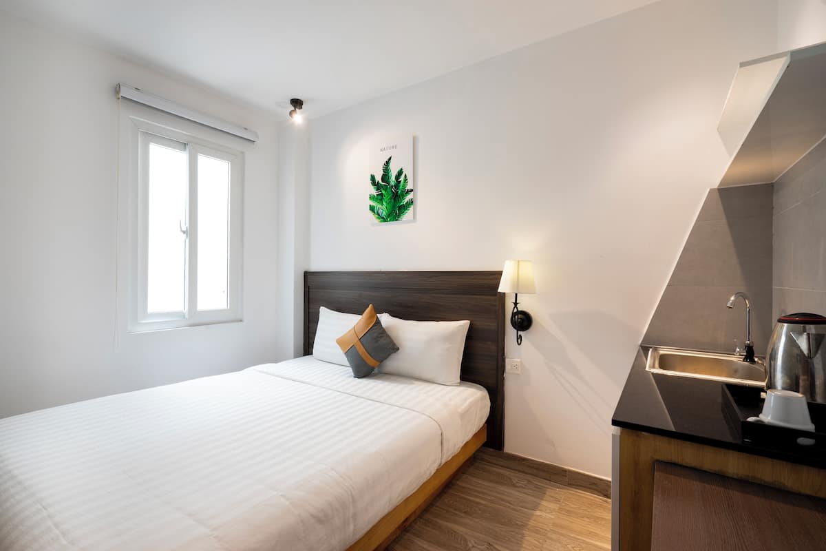 Habitación de hotel con cama, mesita de noche, lámpara de pared, ventana y área de cocina con un lavamanos y un hervidor.