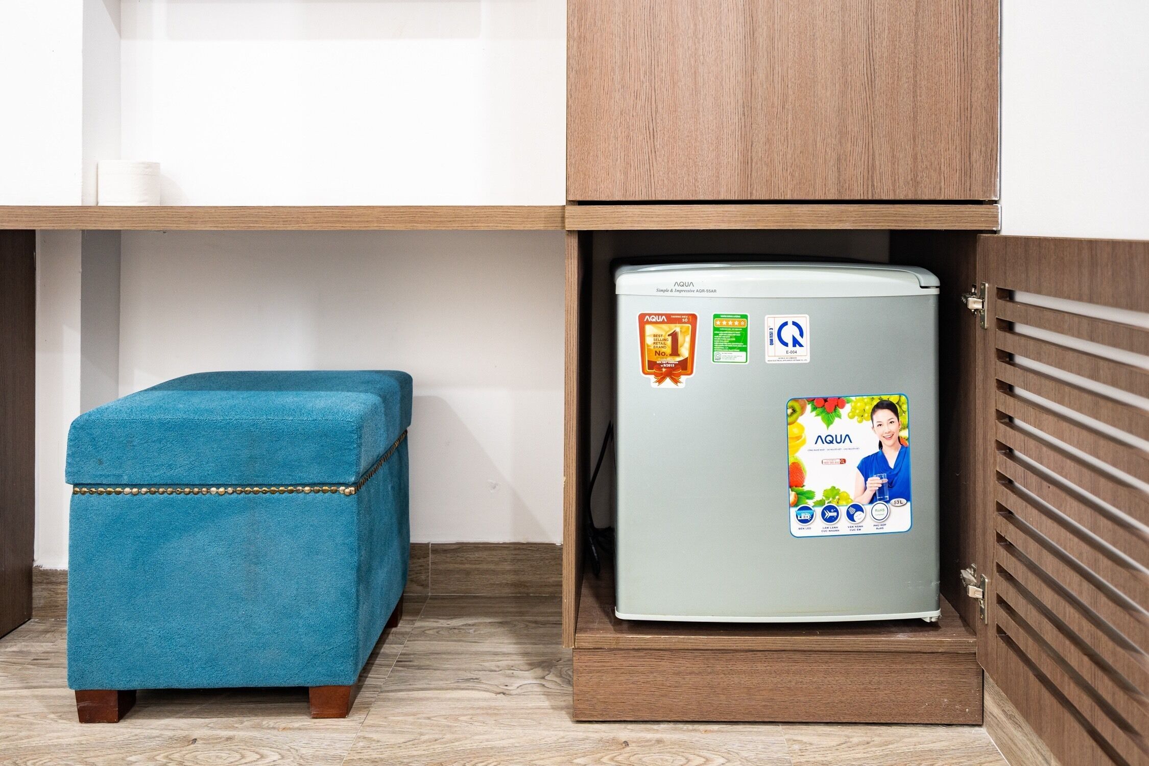 A compact kitchenette with a teal ottoman, a refrigerator with a water bottle advertisement, and wooden cabinetry.