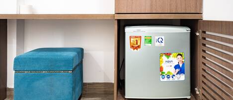 A compact kitchenette with a teal ottoman, a refrigerator with a water bottle advertisement, and wooden cabinetry.