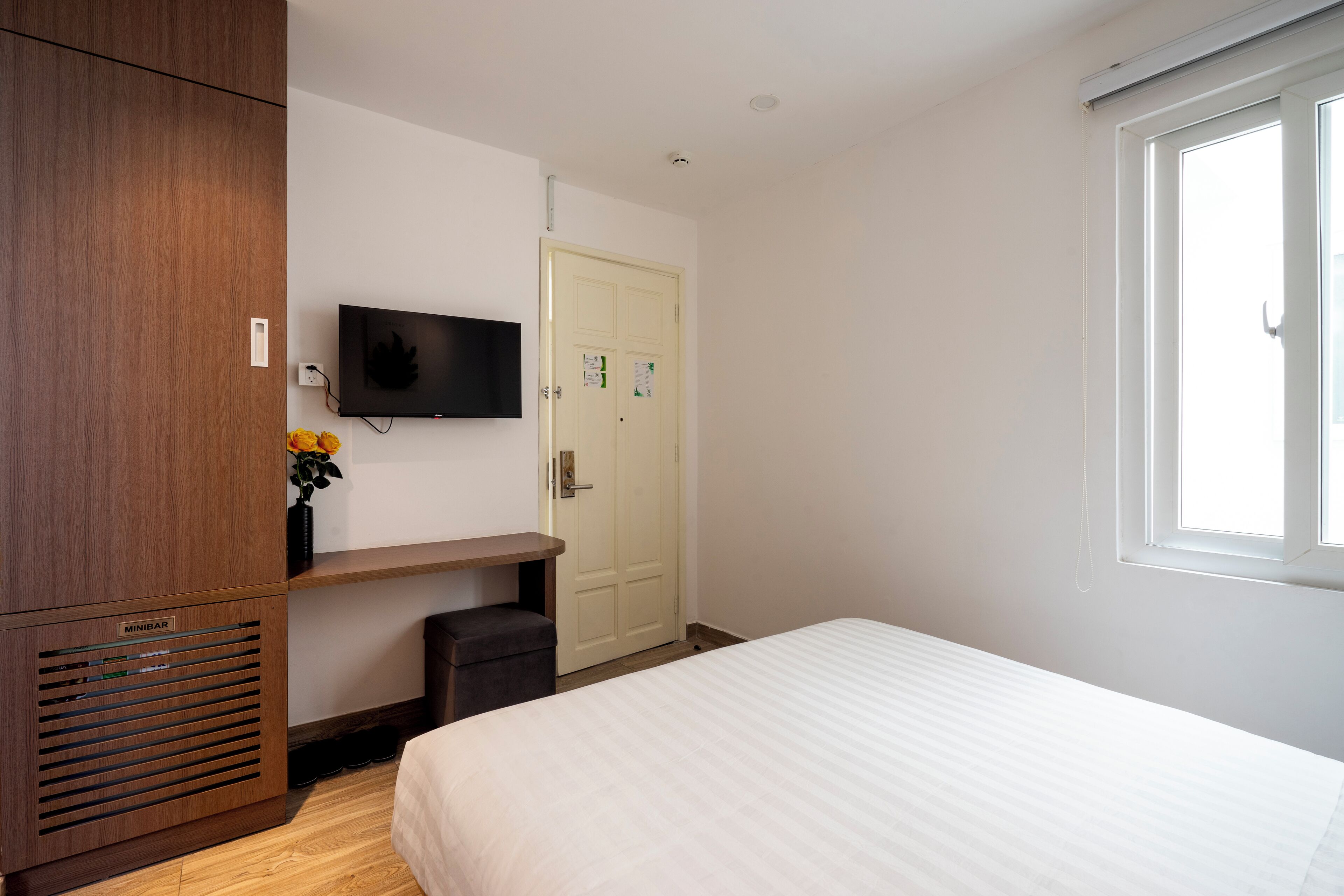 A bedroom with a bed, a TV mounted on the wall, a wooden wardrobe, a small desk with a chair, and a window.