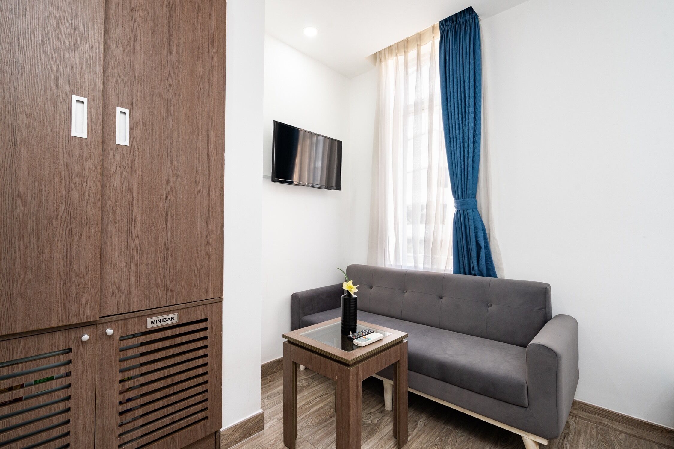 Una habitación moderna con un sofá gris, un armario de madera, una mesita con un jarrón y un control remoto, y un televisor en la pared.