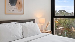 Una cama bien hecha con sábanas blancas, una lámpara de noche y un cuadro abstracto enmarcado en la pared.