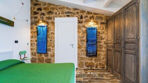 A room with a stone wall, a wooden door, and a green bed. - The Oak and the Donkey. (Piazza Armerina)