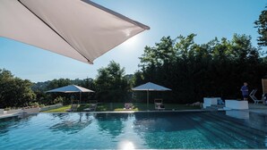 A swimming pool with two sun umbrellas, a clear sky, and a view of trees in the background. - The Oak and the Donkey. (Piazza Armerina)