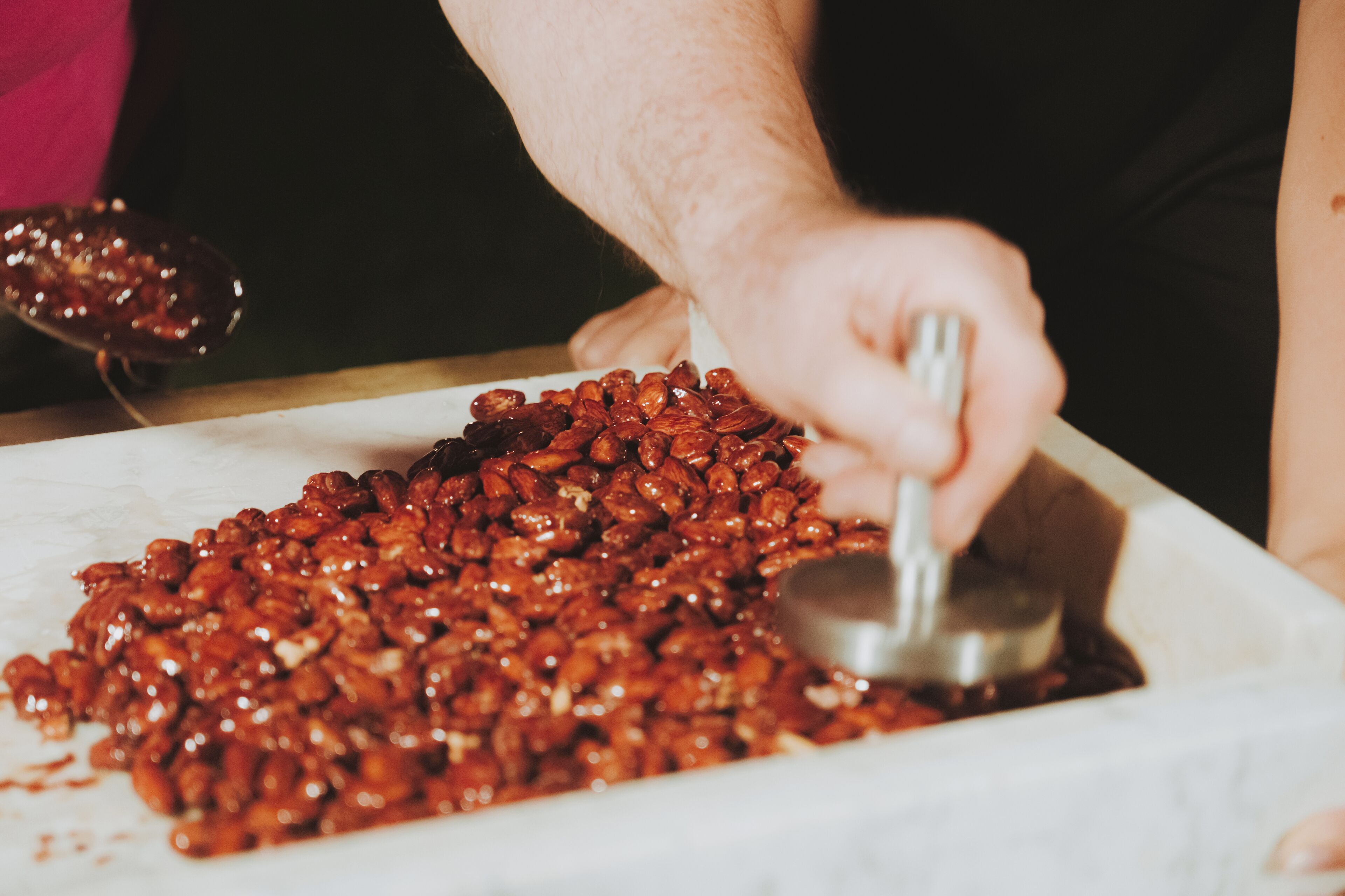 A person scooping a large quantity of glossy, reddish-brown beans from a white container.