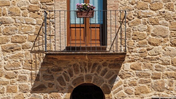 A stone building with a wooden balcony, a window with a flower box, and outdoor seating with chairs and tables. - The Oak and the Donkey. (Piazza Armerina)