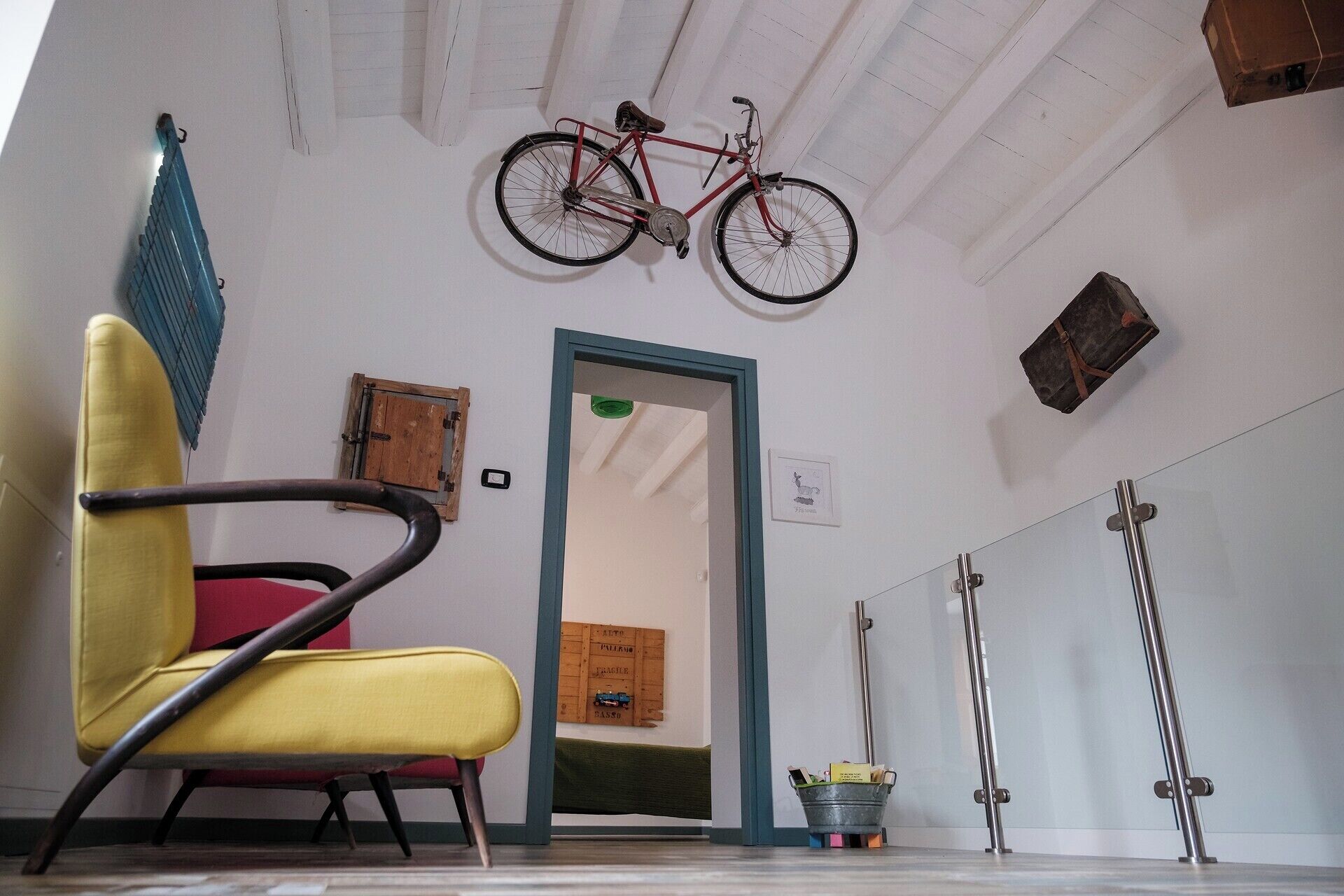 Ein Zimmer mit einem gelben Sessel, einem roten Fahrrad, das von der Decke hängt, einer Holztür und einem Metallregal mit Gegenständen.