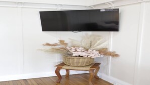 A flat-screen TV mounted on a white wall, a wicker basket with dried plants on a wooden stool below it, and a wooden floor. - Homely 2-Bed Cottage Near Tenterfield Centre (Tenterfield)