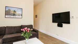Un salon avec un canapé marron, une table basse blanche ornée d’un vase de fleurs, un lustre, un tableau encadré accroché au mur et un téléviseur fixé au mur.