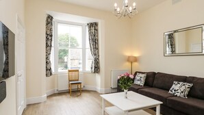Un salon avec un plancher en bois, une table basse blanche, un canapé marron, une chaise en bois, une fenêtre avec des rideaux, une lampe et un miroir au mur.