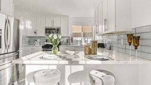 Een moderne keuken met witte kastjes, roestvrijstalen apparatuur, een centraal eiland met barstoelen en uitzicht op de omgeving.