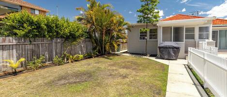 A residential backyard with a wooden fence, a small house, and a covered grill area.