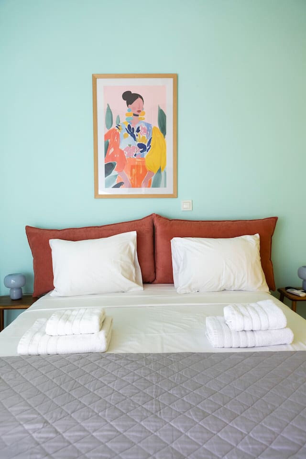 Un lit bien fait, recouvert d’une courtepointe grise matelassée, avec deux oreillers blancs et, au-dessus, un tableau abstrait encadré.