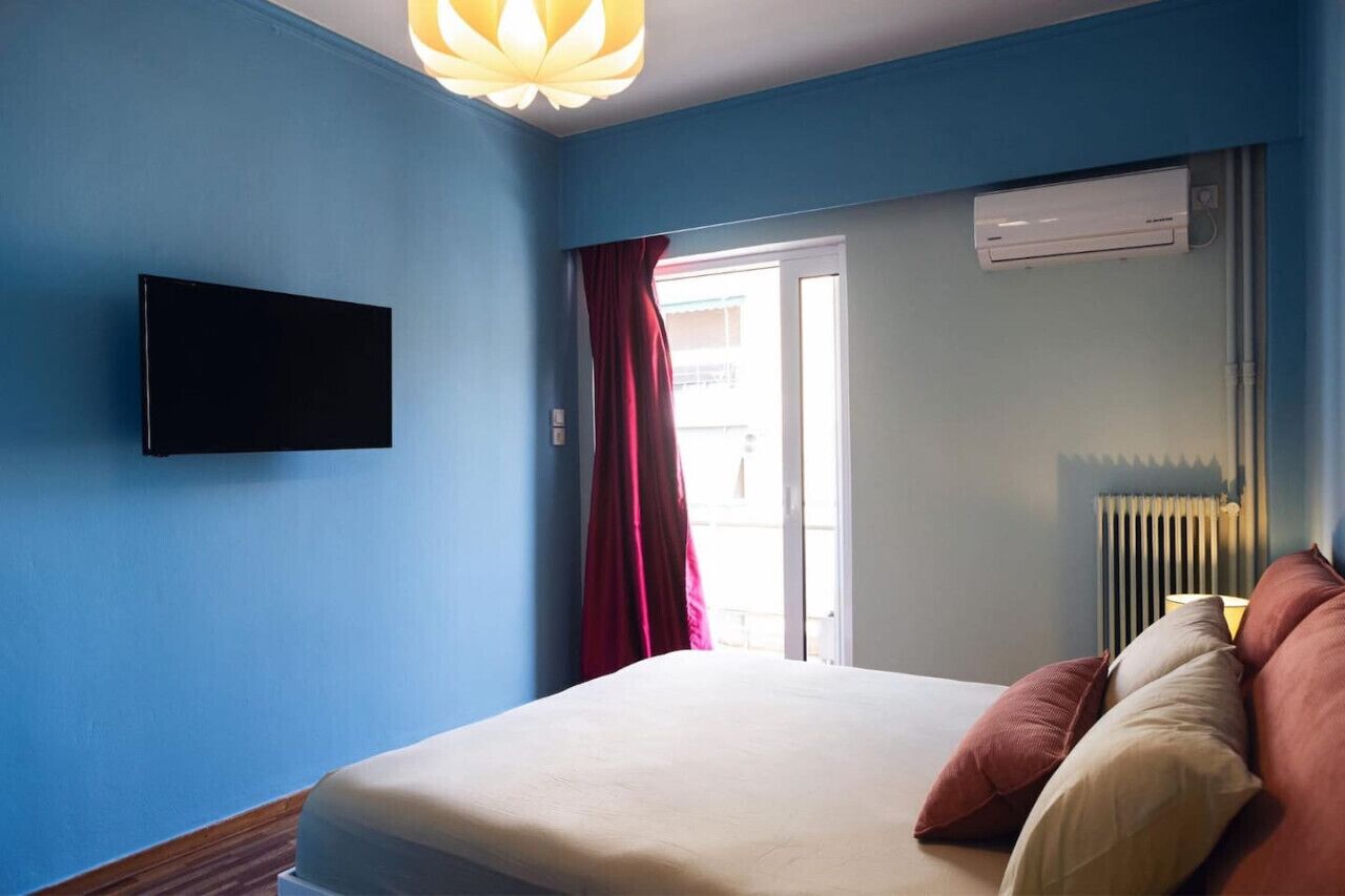 A bedroom with a bed, a flat-screen TV mounted on the wall, a window with a red curtain, and an air conditioning unit.