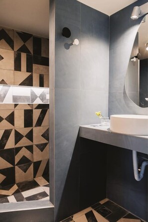 A modern bathroom with a geometric tile pattern on the walls, a white sink, and a mirror above it. - Vibrant Retreat Funky Style & Acropolis View! (Athina)