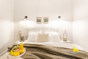 A neatly made bed with white linens and a brown throw blanket, a bedside table with a lamp, a framed picture, and a tray with fruit and juice. - Modern Family Apt with Courtyard near Acropolis! (Athina)