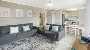 Ein modernes Wohnzimmer mit einem grauen Sofa, einem Couchtisch und einem Teppich. Es gibt eine Küchenzeile mit weißen Schränken und einem Esstisch.