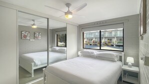 Ein Schlafzimmer mit einem großen Bett, einem Deckenventilator, einem Nachttisch mit Lampe und einem Fenster mit Blick auf Gebäude und Pflanzen.