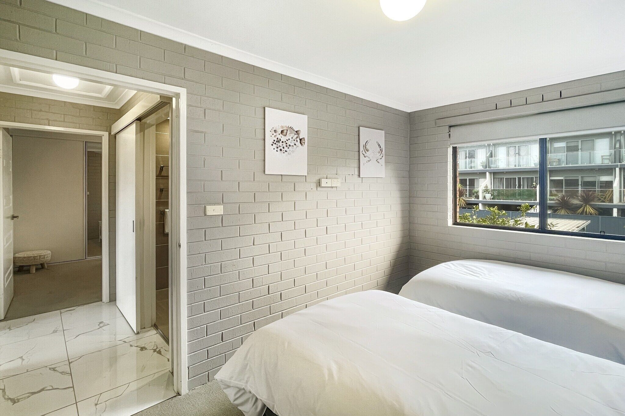 A bedroom with two beds, a window with a view, and a brick wall with artwork.