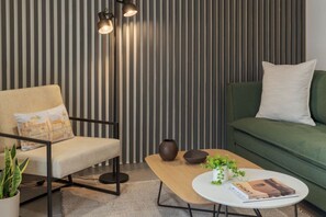 Ein modernes Wohnzimmer mit gestreifter Wand, einem beigefarbenen Sofa, einem grünen Sitzsofa, einem Holztisch und einer Stehlampe.