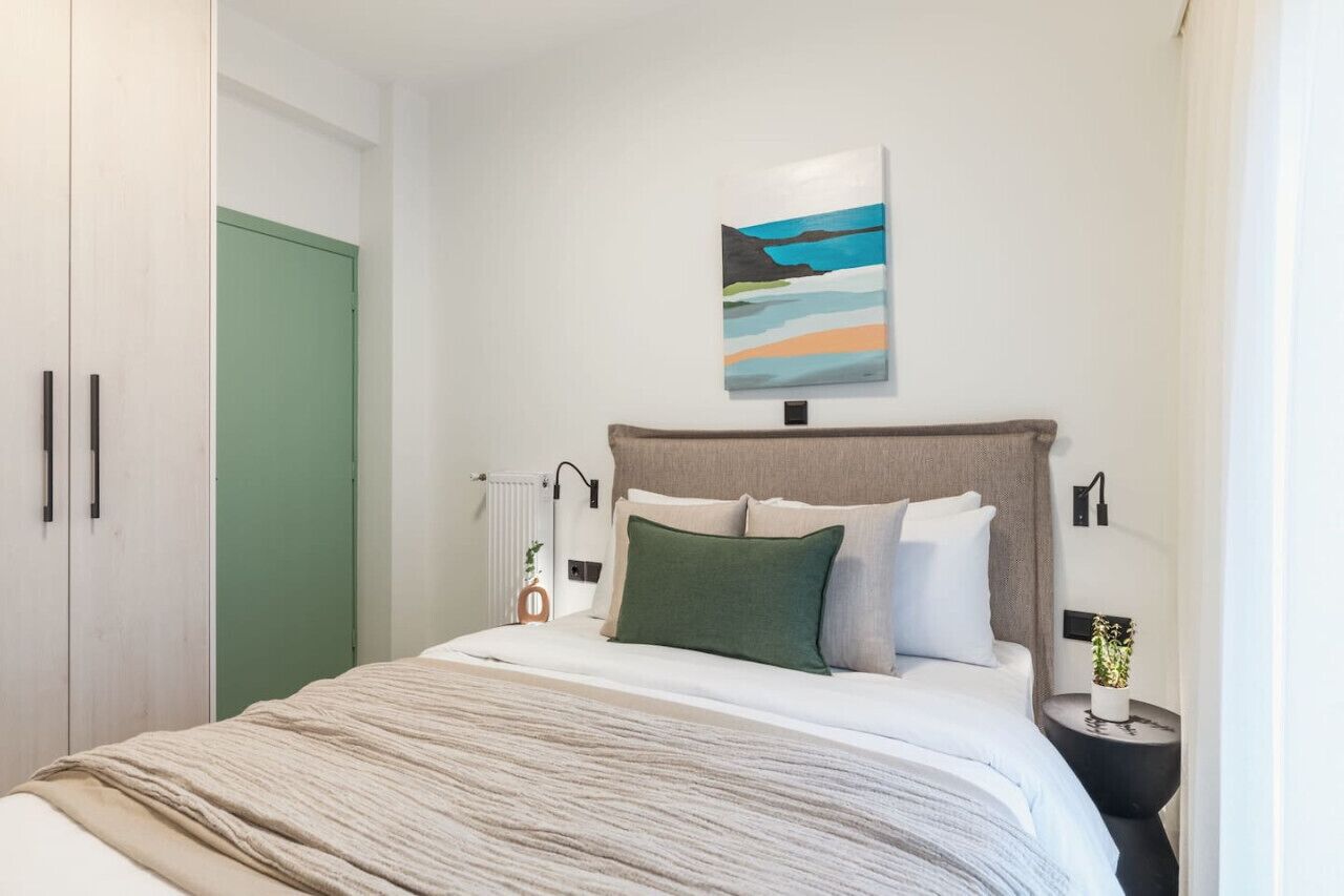 Un dormitorio con una cama, una puerta verde, un cuadro y una mesita de noche con una planta.