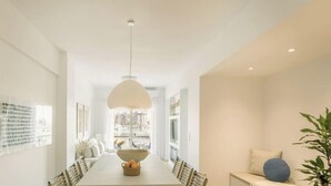Uma área de refeições moderna com uma mesa comprida, cadeiras, um lustre pendente e vista para prédios através de grandes janelas.
