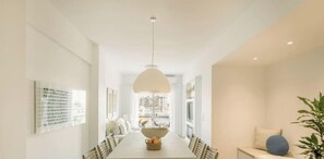 Un espace de restauration moderne avec une longue table, des chaises, un luminaire suspendu et une vue sur les bâtiments à travers de grandes fenêtres.